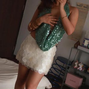 May include: A teal sequined scarf and a white, scalloped mini-skirt. The scarf is draped over the shoulders. Several gold bracelets adorn the arm. The skirt is made of overlapping, circular pieces.