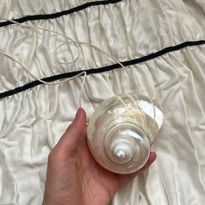 Pode incluir: Uma bolsa de concha perolada com um design em espiral, segurada contra um fundo de tecido branco. A bolsa tem um cordão fino esbranquiçado. O tecido tem uma borda preta.