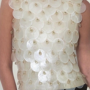 May include: A sleeveless, off-white top adorned with numerous, overlapping, circular, iridescent shell-like elements. Each disc is embellished with a small, dangling, gold-colored accent, creating a shimmering effect. The garment has a scoop neckline.
