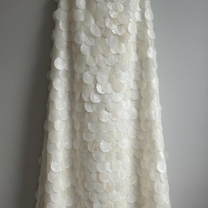 May include: A floor-length, off-white skirt with a unique design. The skirt is covered in overlapping, circular, shell-like embellishments, creating a textured, layered effect. The skirt hangs vertically against a neutral gray background.