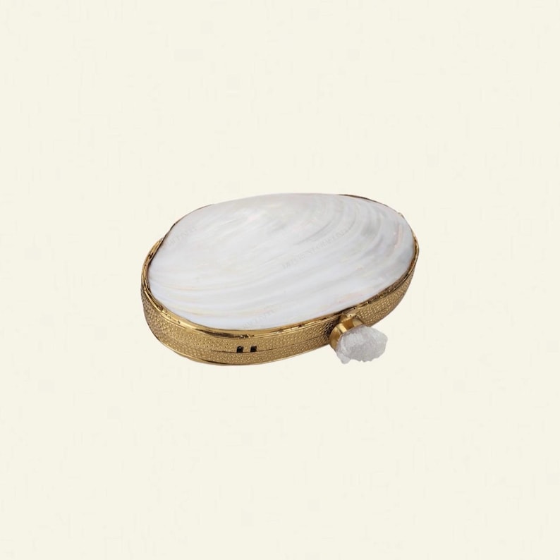 May include: A white clam shell-shaped clutch with a gold-coloured metal frame. The clutch features a textured gold band and a small, white crystal accent. The shell has a pearlescent sheen.