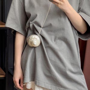 May include: A grey oversized t-shirt with a white seashell purse. The purse is attached to a thin, white cord. The bottom of the shirt is paired with a white lace trim.