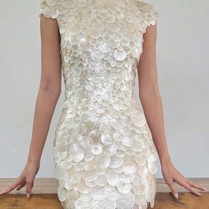 May include: A cream-colored, short-sleeved dress covered in overlapping, circular, shell-like pieces. The dress has a fitted bodice and a short skirt. The model is standing with arms at their sides, hands resting on a wooden surface.
