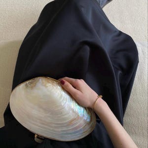 May include: A large, iridescent seashell clutch with a gold-toned trim. The clutch is held by a person wearing a black dress and a gold bracelet. The shell has a pearlescent sheen with hints of blue and green.