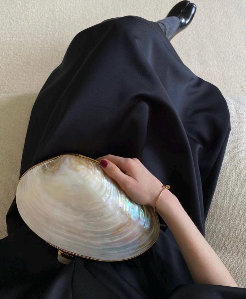 May include: A large, iridescent seashell clutch with a gold-toned trim. The clutch is held by a person wearing a black dress and a gold bracelet. The shell has a pearlescent sheen with hints of blue and green.