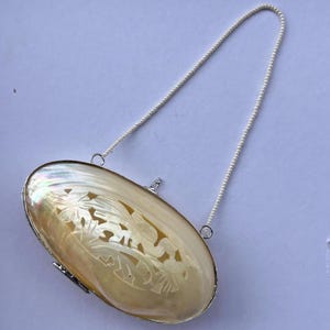 Könnte beinhalten: Ovale, schillernde Perlmutt-Clutch mit silberfarbenem Rahmen und zartem Perlenhenkel. Die Clutch hat ein geschnitztes Blumendesign. Ein kleines, elegantes Accessoire.