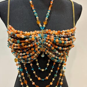 May include: A beaded top with orange, teal, and gold beads. The top is made of various sizes of beads and has gold chain straps. The design includes cascading bead strands and a central pendant. The top is displayed on a black mannequin.