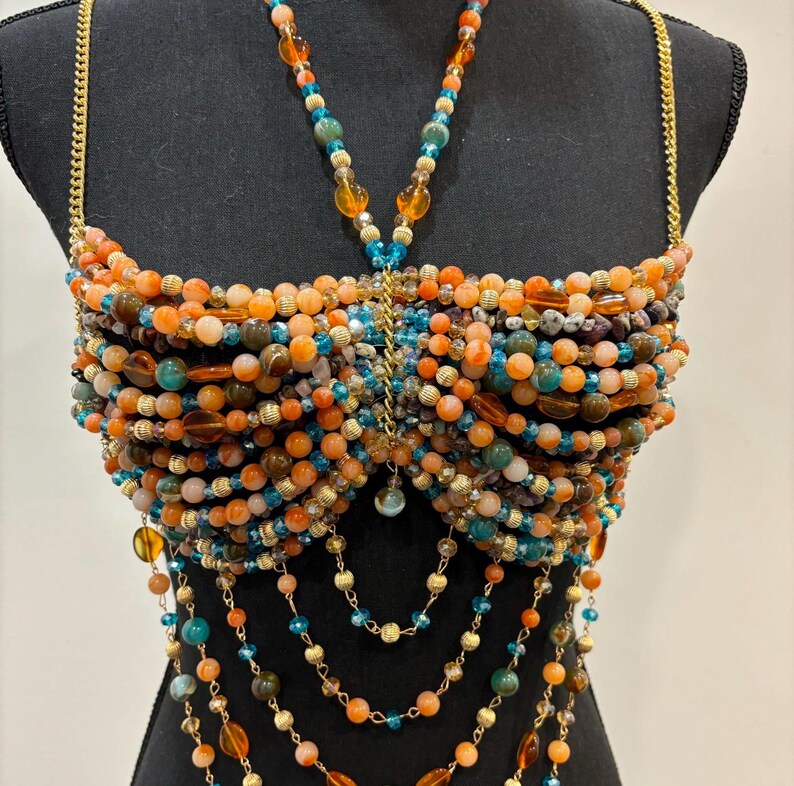 May include: A beaded top with orange, teal, and gold beads. The top is made of various sizes of beads and has gold chain straps. The design includes cascading bead strands and a central pendant. The top is displayed on a black mannequin.