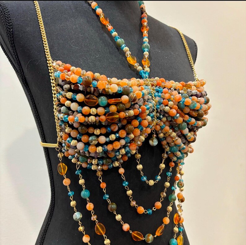 May include: A beaded top in shades of orange, brown, and teal, displayed on a black mannequin. The top is constructed with rows of beads and gold-colored chain straps. Dangling strands of beads cascade from the bottom.