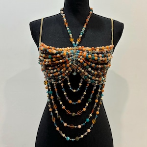 May include: A beaded top in shades of orange, teal, and brown, displayed on a black mannequin. The top features a halter-style neckline and cascading beaded strands. The beads are various sizes and shapes, creating a textured and layered effect. Gold chain straps.