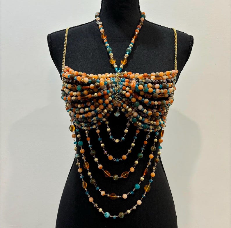 May include: A beaded top in shades of orange, teal, and brown, displayed on a black mannequin. The top features a halter-style neckline and cascading beaded strands. The beads are various sizes and shapes, creating a textured and layered effect. Gold chain straps.