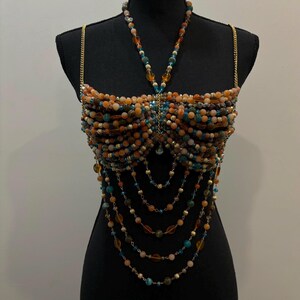 May include: A beaded top in shades of orange, brown, and teal, displayed on a black mannequin. The top features a halter-style neckline and gold chain straps. Long beaded strands cascade down from the bust, creating a layered effect. The beads are various sizes and shapes.