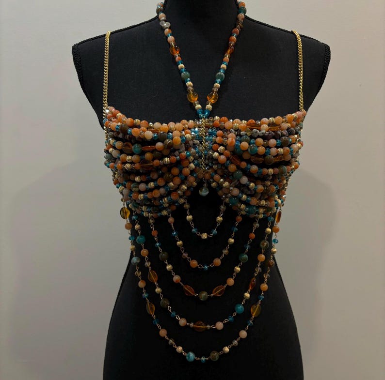 May include: A beaded top in shades of orange, brown, and teal, displayed on a black mannequin. The top features a halter-style neckline and gold chain straps. Long beaded strands cascade down from the bust, creating a layered effect. The beads are various sizes and shapes.