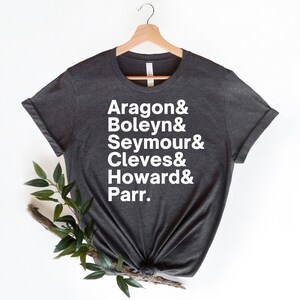 May include: A dark grey t-shirt with white text that reads "Aragon & Boleyn & Seymour & Cleves & Howard & Parr."
