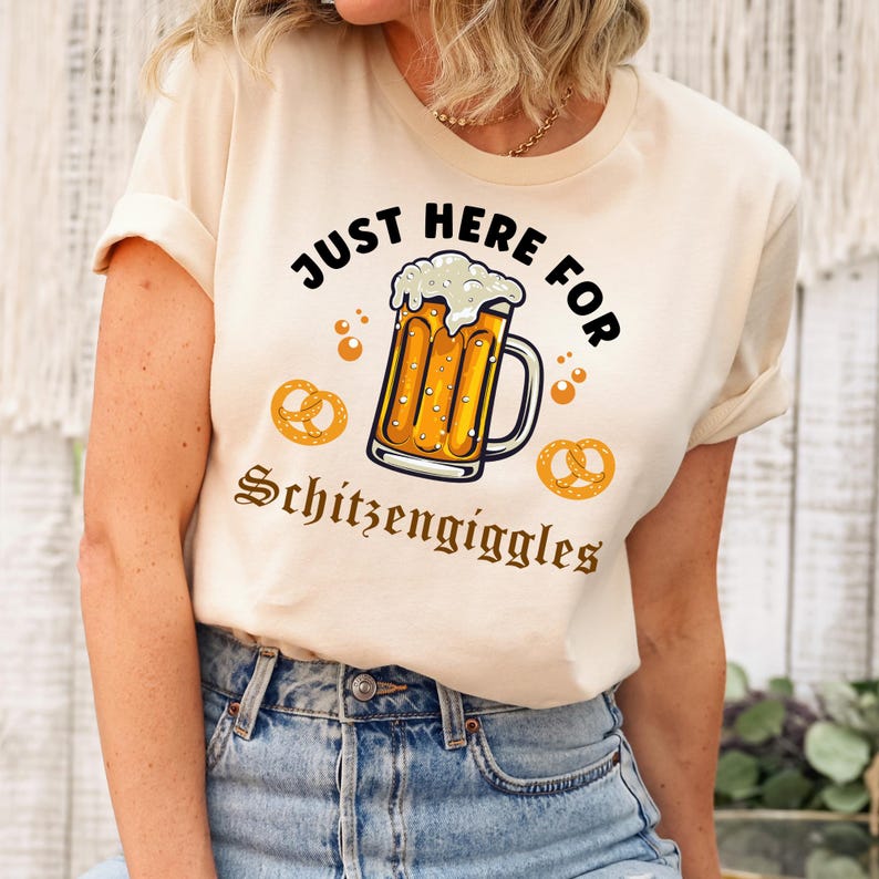 Op de afbeelding: Een beige T-shirt met de tekst "Just Here For Schitzen giggles" in bruine letters. Boven de tekst staat een cartoonillustratie van een bierglas met schuim en pretzels.