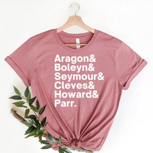 May include: A dusty rose t-shirt with white text that reads "Aragon & Boleyn & Seymour & Cleves & Howard & Parr."