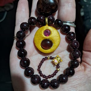 May include: A bracelet made of dark red, spherical beads, a circular yellow pendant featuring a small pink stone and a larger dark red stone, and a smaller ring with tiny dark red beads and a gold-toned bear charm with a pink heart accent.
