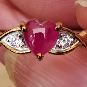 May include: A gold ring featuring a central heart-shaped cabochon ruby gemstone. The ruby is flanked on each side by a small, clear, round-cut diamond set within a silver-toned triangular accent. The band is polished gold with a smooth, tapered design.
