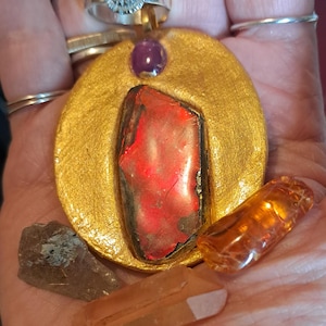May include: A gold-colored pendant with a large red stone and a small purple stone. Several other gemstones are also present, including a clear orange crystal and a smoky quartz. A silver ring with a red stone is also visible.