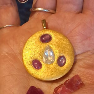 May include: A circular gold-toned pendant featuring a central pear-shaped clear stone surrounded by three oval purple cabochon stones. A silver-toned ring with a purple stone and a plain silver-toned band are visible, along with two loose red stones.