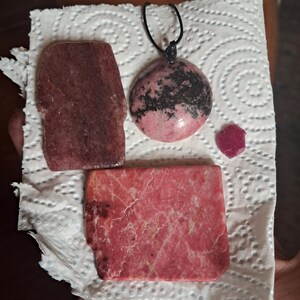May include: A collection of pink and red stones including a rectangular slab, a smaller irregular slab, a round pendant on a black cord with black inclusions, and a small, bright red, angular crystal fragment with a textured surface.