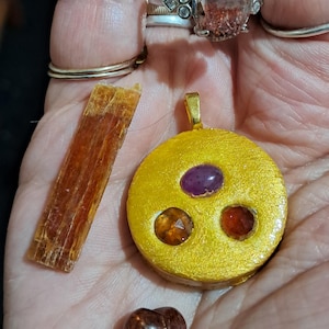 Könnte beinhalten: Eine Sammlung von Schmuck und Edelsteinen, darunter ein goldener Anhänger mit drei eingesetzten Steinen, ein herzförmiger Stein, ein rechteckiger Stein und Ringe. Der Anhänger hat eine goldene Oberfläche und verfügt über einen lila, orangefarbenen und roten Stein.