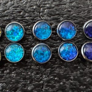 May include: A selection of stud earrings with a silver-coloured metal setting. The earrings display a range of colours, including red, yellow, green, blue, purple, and orange. The earrings are arranged in two rows on a dark textured surface.