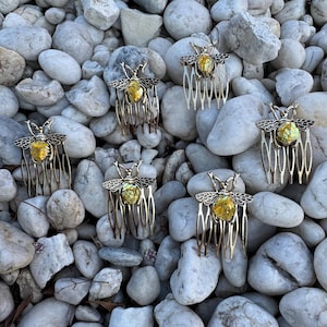 May include: Six gold bee hair combs with yellow and green iridescent wings and bodies. The combs are on a bed of white stones.