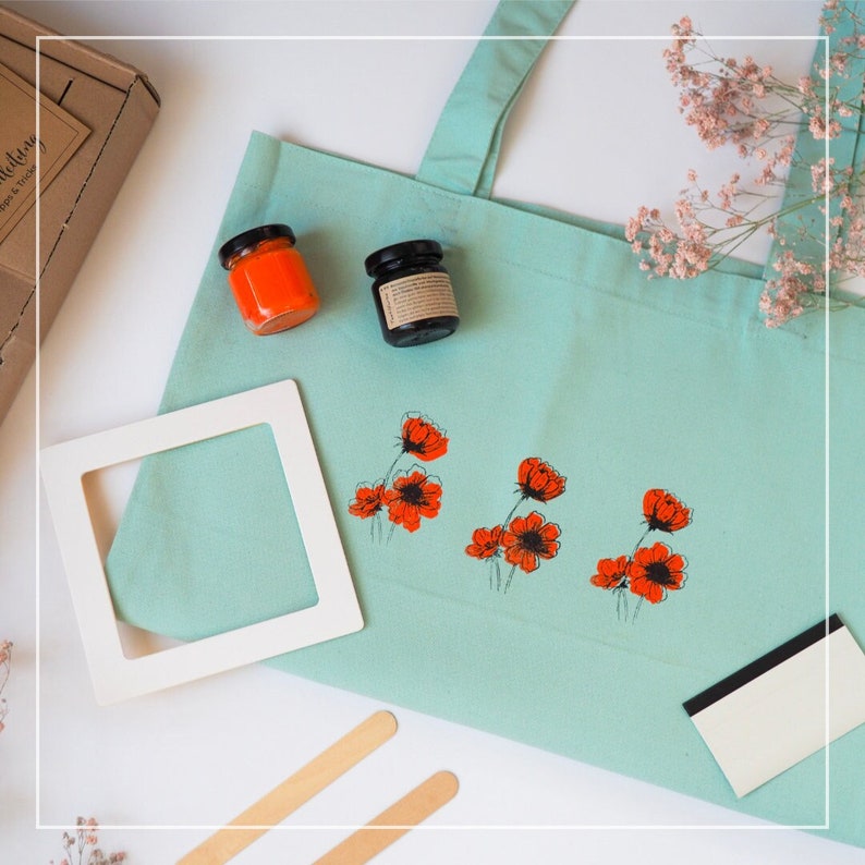 May include: A teal tote bag with a white frame, two small jars of orange paint, a black jar of paint, two wooden craft sticks, a white squeegee, and a design of red poppy flowers printed on the bag.