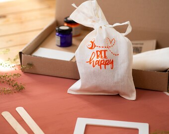 May include: A white canvas bag with a drawstring closure and the text "bee happy" printed in orange. The bag is sitting on a wooden surface with a box of craft supplies in the background.