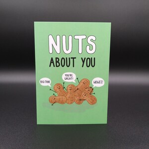 May include: A green card with the text "NUTS ABOUT YOU" in white.  The card features a cartoon illustration of peanuts with faces and arms, with speech bubbles that say "BIG FAN!" "YOU'RE GREAT!" and "WOWEE!"