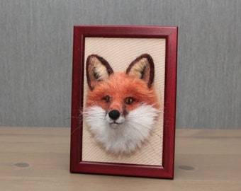 May include: A framed artwork featuring a detailed, three-dimensional fox head. The fox has orange fur, dark brown ears, a black nose, and white fur around its muzzle. The artwork is set in a dark red wooden frame.