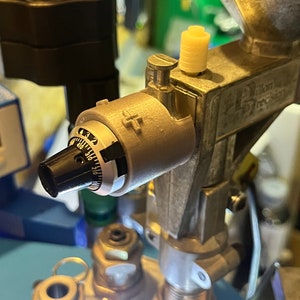 Op de afbeelding: Close-up van een zilveren metalen apparaat met een zwarte knop en een wijzerplaat die "2 tot 6" aangeeft. Het apparaat heeft een witte kunststof knop en een zilveren metalen hendel. Het apparaat is bevestigd aan een groter zilveren metalen apparaat met een trechter aan de bovenkant.