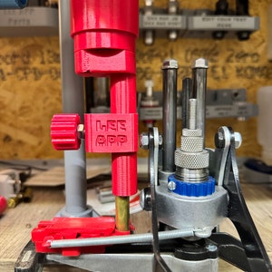 Può includere: Una pressa di ricarica rossa con una base argentata e blu. La pressa ha una leva e un manico. La pressa è montata su una superficie di legno. La pressa ha un'etichetta che dice "LEE APP".