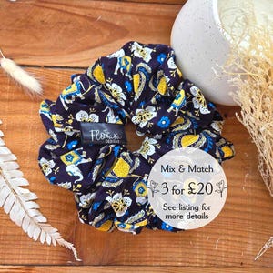 May include: A dark blue and yellow patterned hair scrunchie with white floral accents. The scrunchie has the text "Flotare Designs" on it. The text "Mix & Match 3 for £20 See listing for more details" is on a white circle.