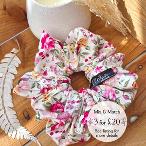 May include: A white scrunchie with a floral print in shades of pink, yellow, and green. The scrunchie has a black tag with the text "Flotara Designs". The text "Mix & Match 3 for £20. See listing for more details" is also visible in the image.