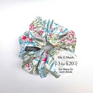 May include: A white scrunchie with a floral print featuring pink, blue, and green flowers. The scrunchie has a tag that says "Flora Designs". The text "Mix & Match 3 for £20 See listing for more details" is also visible.
