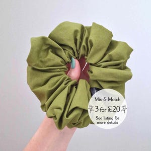 May include: An olive green fabric scrunchie. The scrunchie is held in a hand. The image includes the text 'Mix & Match 3 for £20'. A hair accessory.