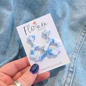 May include: Cloud-shaped earrings in blue and white, with silver-coloured posts. The earrings are presented on a white card with the text "Floara Designs" and the website address www.floara.co.uk.
