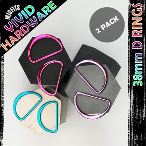 May include: A selection of D-rings in shades of pink and teal, presented on geometric forms. The image includes text that reads "Vivid Hardware" and "38mm D Rings". The package is labeled "2 Pack".