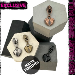 May include: Four zipper pulls with geometric heart-shaped charms in matte finishes: black, silver, gold, and copper. The text "GEO HEART ZIPPER PULL" and "EXCLUSIVE" are visible in the image. The text "MATTE FINISHES" is also visible.