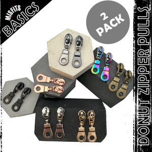 May include: Two packs of donut zipper pulls in black, silver, rose gold, and rainbow colours. The zipper pulls are in the shape of a donut. The image text reads "Misfits Basics 2 Pack Donut Zipper Pulls".