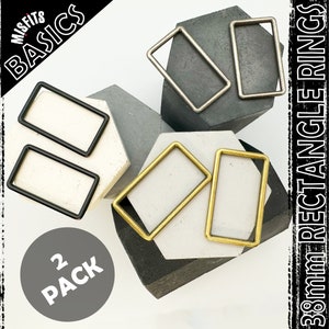 Puede incluir: Dos paquetes de anillos rectangulares de 38 mm en negro, plata y oro. Los anillos se muestran en superficies grises y negras. La imagen también incluye el texto "Misfits Basics" y "2 Pack".