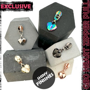 以下が含まれることがあります： 虹色、シルバー、ゴールド、ローズゴールドの4種類の光沢仕上げの幾何学的なハート型のジッパー引き手。ジッパー引き手は小さなコンクリートブロックの上に置かれています。画像には「GEO HEART ZIPPER PULL」と「EXCLUSIVE」のテキストが表示されています。