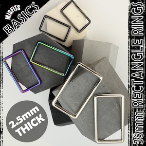 Puede incluir: Cinco anillos metálicos rectangulares de diferentes tamaños y acabados, incluyendo plata, negro e iridiscente. Los anillos se muestran sobre un fondo de hormigón gris. El texto "Misfits Basics" y "38mm Rectangle Rings" es visible en el fondo. El texto "2.5mm Thick" es visible en primer plano.