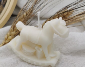 May include: White candle shaped like a rocking horse. The candle is on a white fabric background.