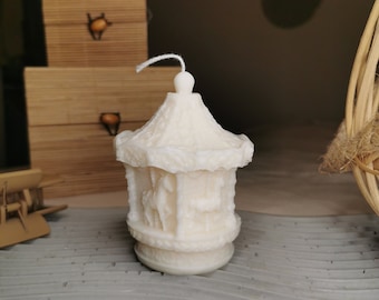 May include: Ivory-colored carousel candle with detailed relief carvings of horses and decorative elements. The candle has a domed roof and a central wick. The candle is placed on a gray, textured surface.