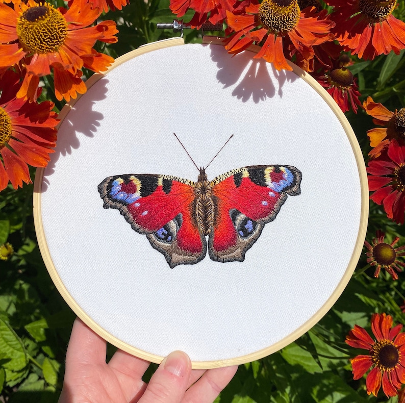 May include: Embroidered peacock butterfly on white fabric in a wooden embroidery hoop. The butterfly is red, brown, and blue.