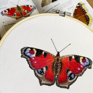 May include: Embroidered peacock butterfly with red, brown, and blue wings. The butterfly is stitched on white fabric in a circular embroidery hoop.