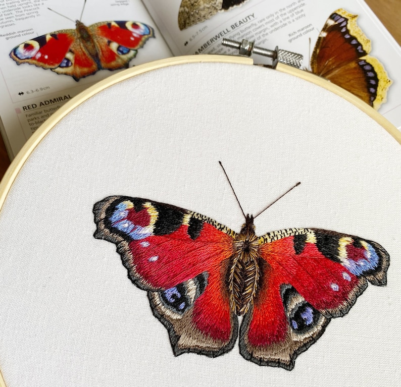 May include: Embroidered peacock butterfly with red, brown, and blue wings. The butterfly is stitched on white fabric in a circular embroidery hoop.